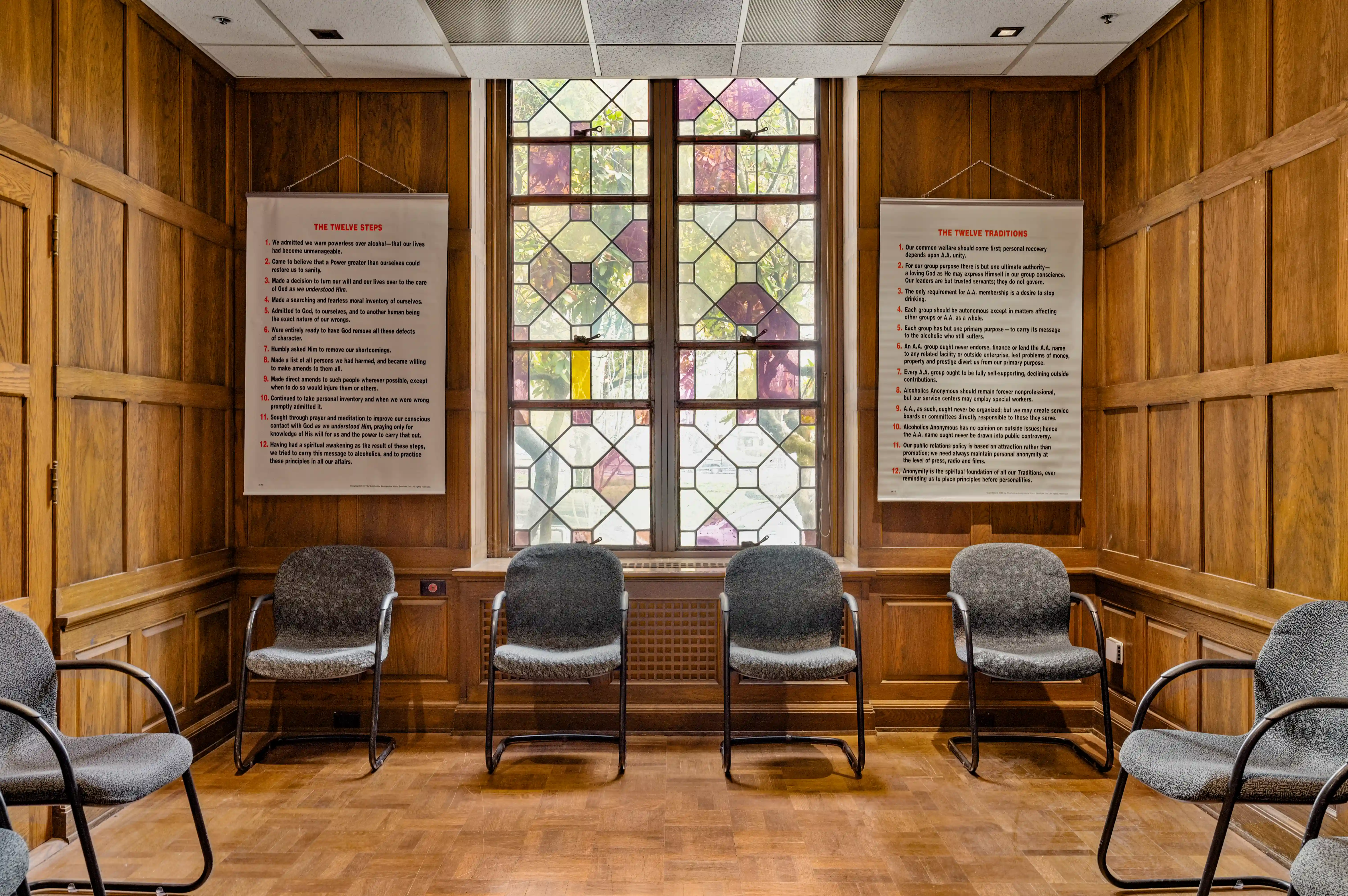 Group therapy room with Twelve Steps and Twelve Traditions posters flanking stained glass window at Atlas Treatment Center